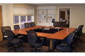 Interior meeting room with U-shaped wooden tables surrounded by office chairs, shelving, and windows.