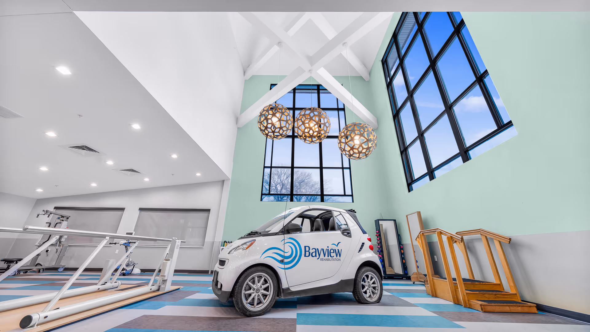 Interior rehabilitation gym with a Bayview-branded small car parked on a patterned floor beneath large windows and hanging pendant lights.