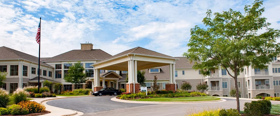 Exterior view of Wesley Willows senior living facility showing a large building with multiple windows, a covered entrance with columns, landscaped greenery, a tree, and an American flag on a flagpole under a partly cloudy sky.