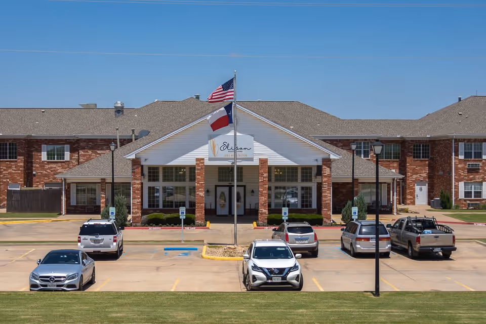 Front exterior view of Elison Assisted Living of Lake Wellington building with a parking lot in front. The building is made of red brick with a white entrance featuring two flags, the American flag and the Texas state flag, flying on flagpoles. Several cars are parked in the lot under a clear blue sky.