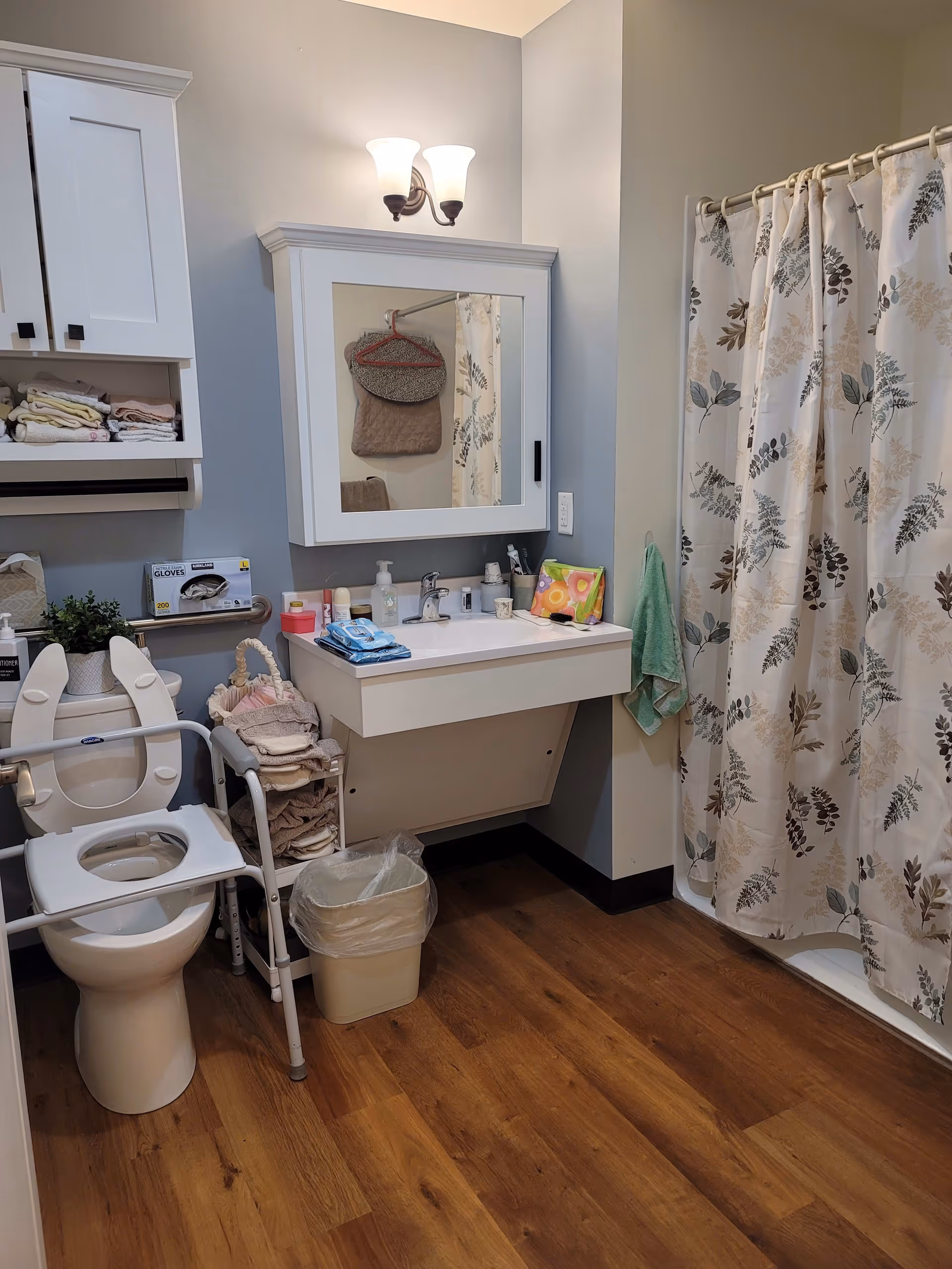 A bathroom with a toilet equipped with safety rails, a white sink with various toiletries on the counter, a mirror with a cabinet above the sink, a shower with a floral-patterned curtain, wooden flooring, and a small trash bin. Towels and gloves are also visible on shelves and racks.