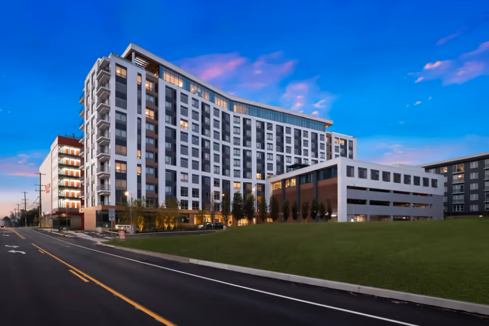 Exterior view of a modern multi-story senior living facility building named Revelle King of Prussia at dusk, with lights on inside and a clear sky with some clouds.