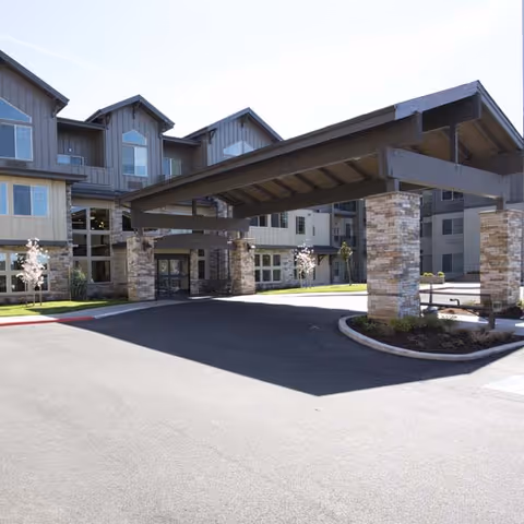 Exterior view of a senior living facility with a large covered entrance supported by stone pillars, surrounded by a paved driveway and landscaped areas with small trees and shrubs.