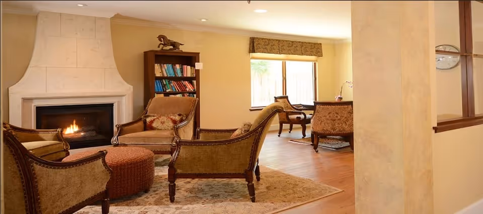 A cozy living room with a lit fireplace, four upholstered armchairs arranged around a round ottoman on a patterned rug, wooden flooring, and a window with a valance letting in natural light.