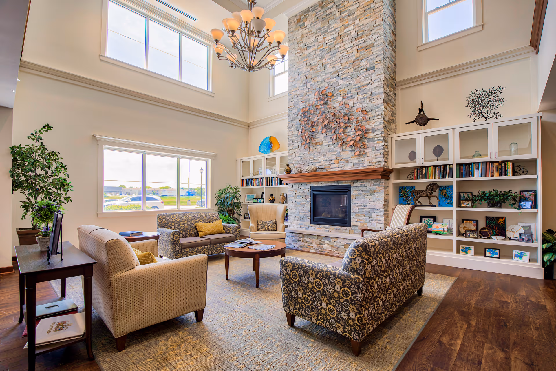 A bright and spacious living room area with high ceilings and large windows. The room features a stone fireplace with decorative art above it, surrounded by built-in white shelves filled with books and decorative items. There are multiple upholstered chairs and sofas arranged around a wooden coffee table on a patterned rug. A chandelier hangs from the ceiling, and there are several green plants adding a touch of nature to the space.