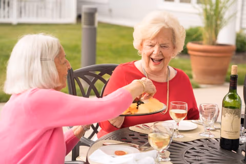 Two elderly women sitting outdoors at a round metal table, sharing a plate of food. One woman in a pink sweater is passing the plate to the other woman in a red top who is smiling. The table has wine glasses, a bottle of wine, and placemats. There is greenery and a potted plant in the background.
