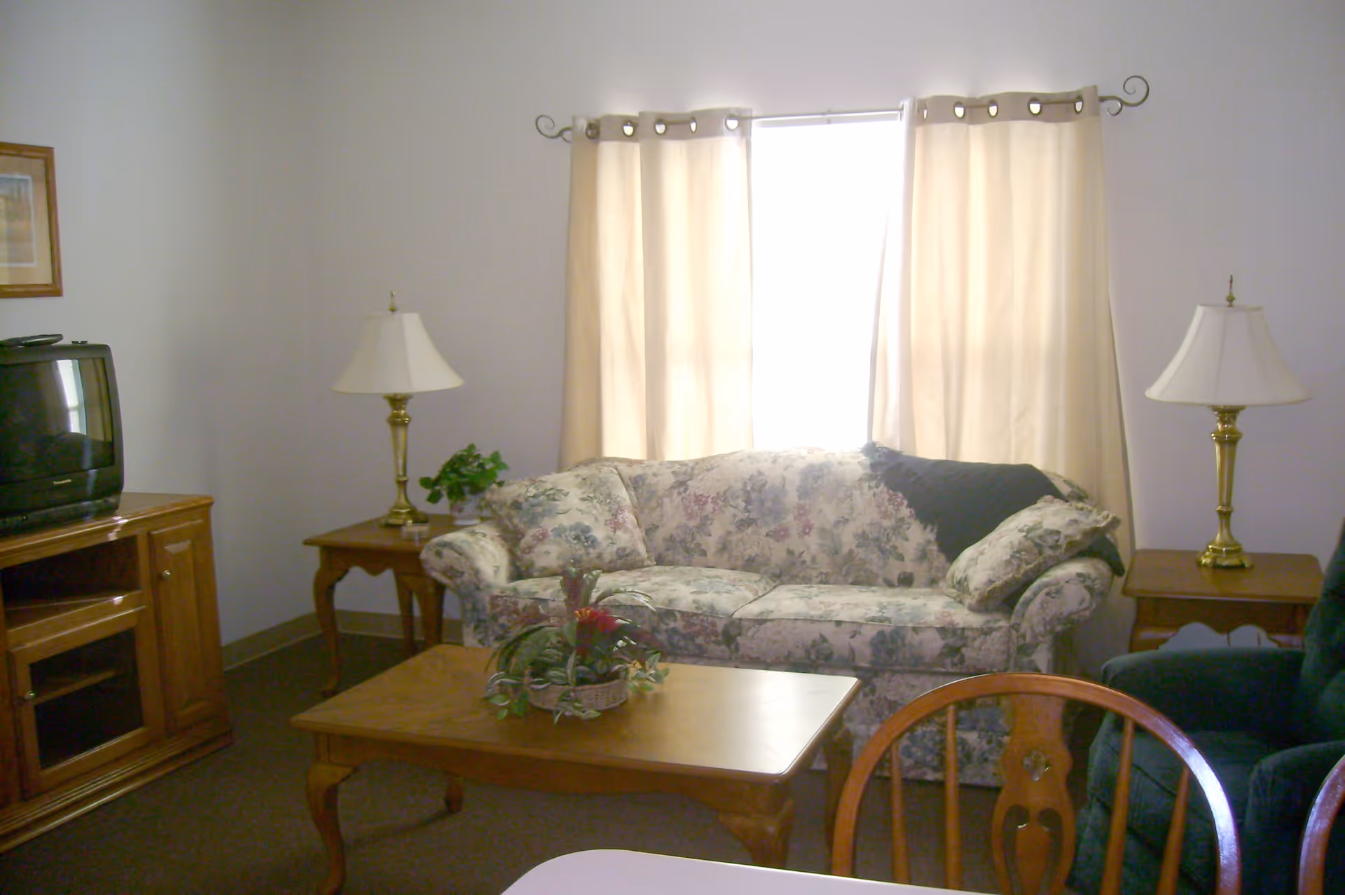 A cozy living room with a floral patterned sofa, two wooden side tables each with a brass lamp, a wooden coffee table with a plant centerpiece, a dark green armchair, a wooden TV stand with an older model television, and beige curtains covering a window.