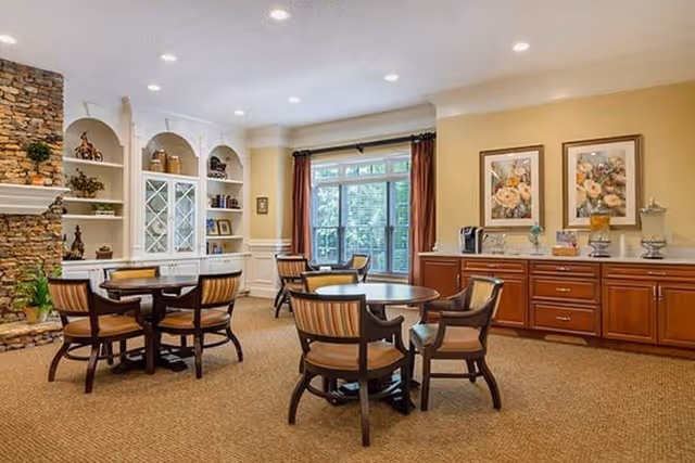 A cozy common area with round wooden tables and cushioned chairs arranged on a carpeted floor. The room features a stone fireplace on the left, built-in white shelves with decorative items, large windows with brown curtains letting in natural light, and a wooden cabinet with framed floral artwork above it on the right.