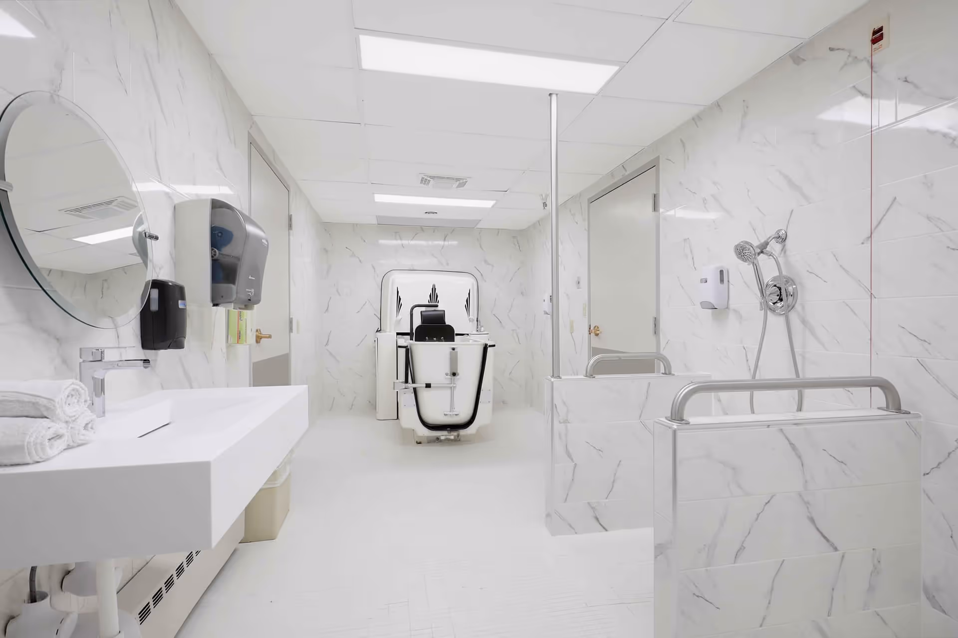 Bright white hospital bathroom with marble walls, sink, mirror, and a chair-looking device at the back.