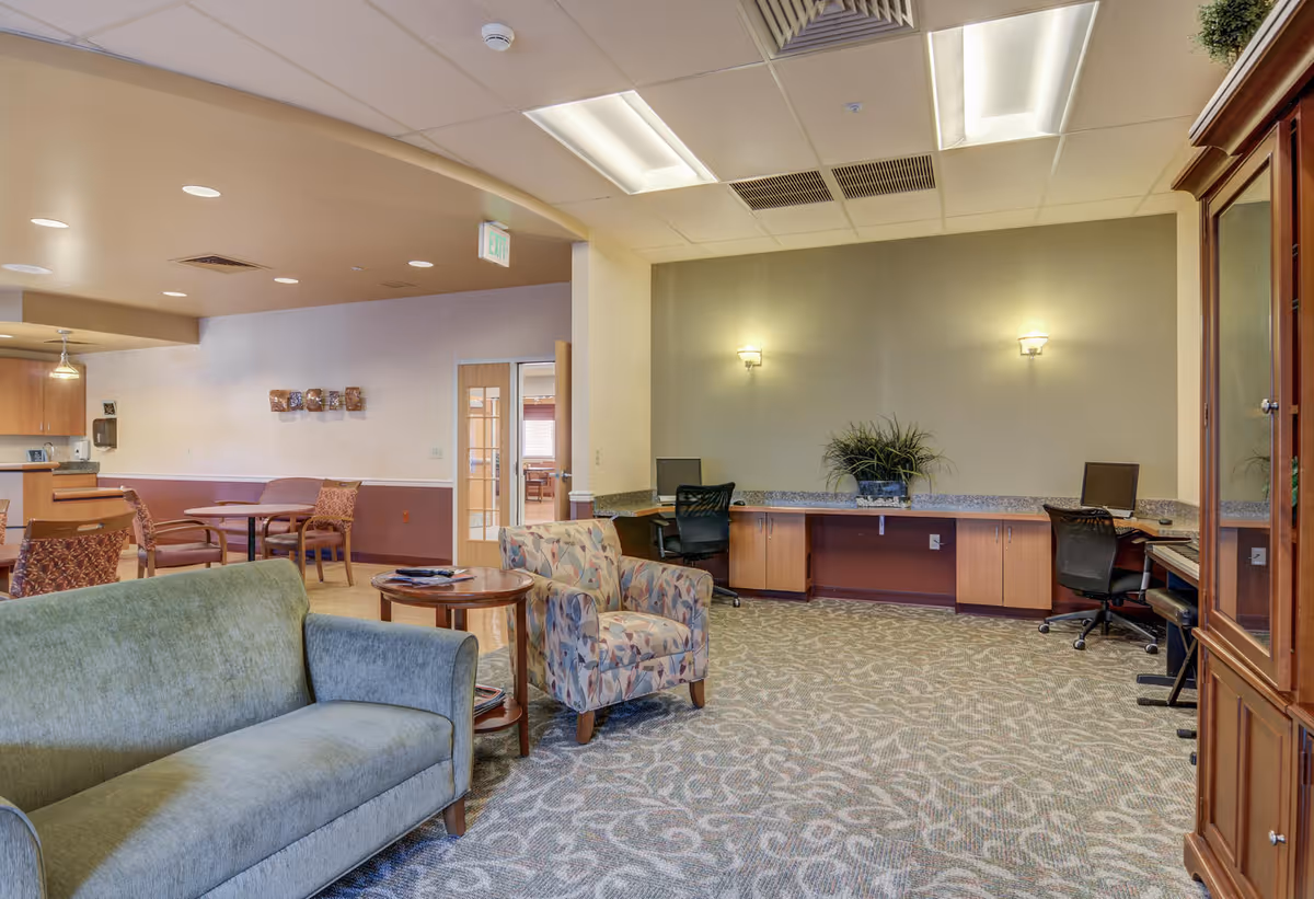 A spacious common area in a senior living facility featuring a green sofa, a patterned armchair, a round wooden side table, and a long counter with two computer workstations and office chairs. The room has carpeted flooring with a decorative pattern, soft wall lighting, and an open view into a dining area with tables and chairs.