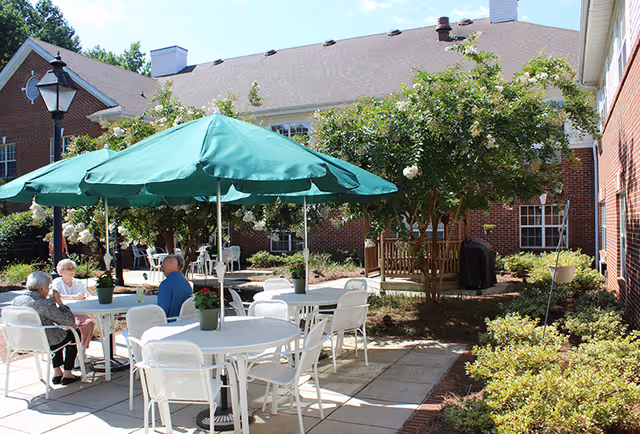 Outdoor patio area at Carriage House Senior Living Community with white tables and chairs under green umbrellas. Several seniors are seated at the tables enjoying the sunny day. The patio is surrounded by well-maintained bushes, trees, and a brick building in the background.