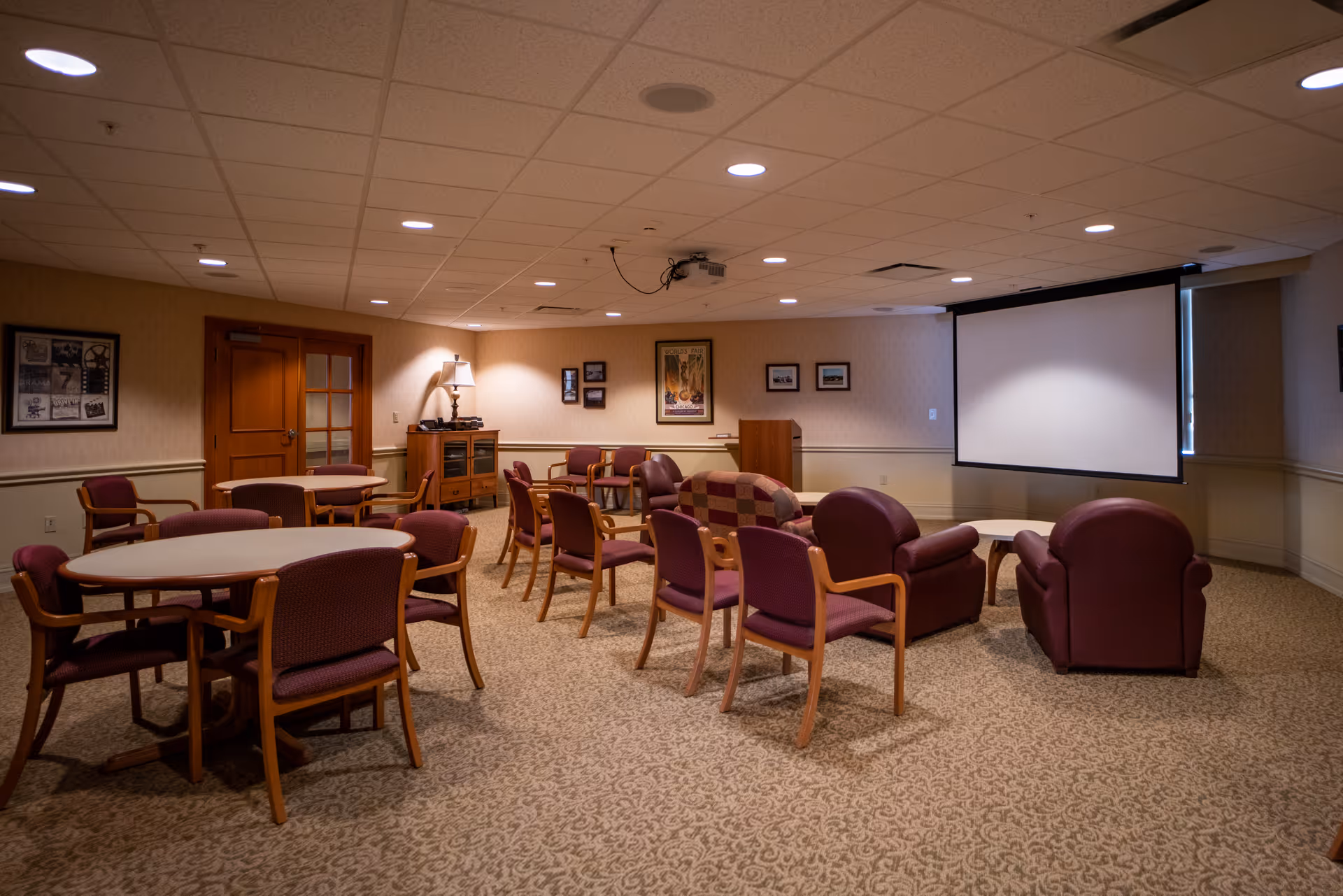 A community room with multiple round tables and chairs arranged for group seating. There are also several armchairs and a couch facing a large projection screen on the wall. The room has carpeted floors, framed pictures on the walls, and a ceiling-mounted projector.