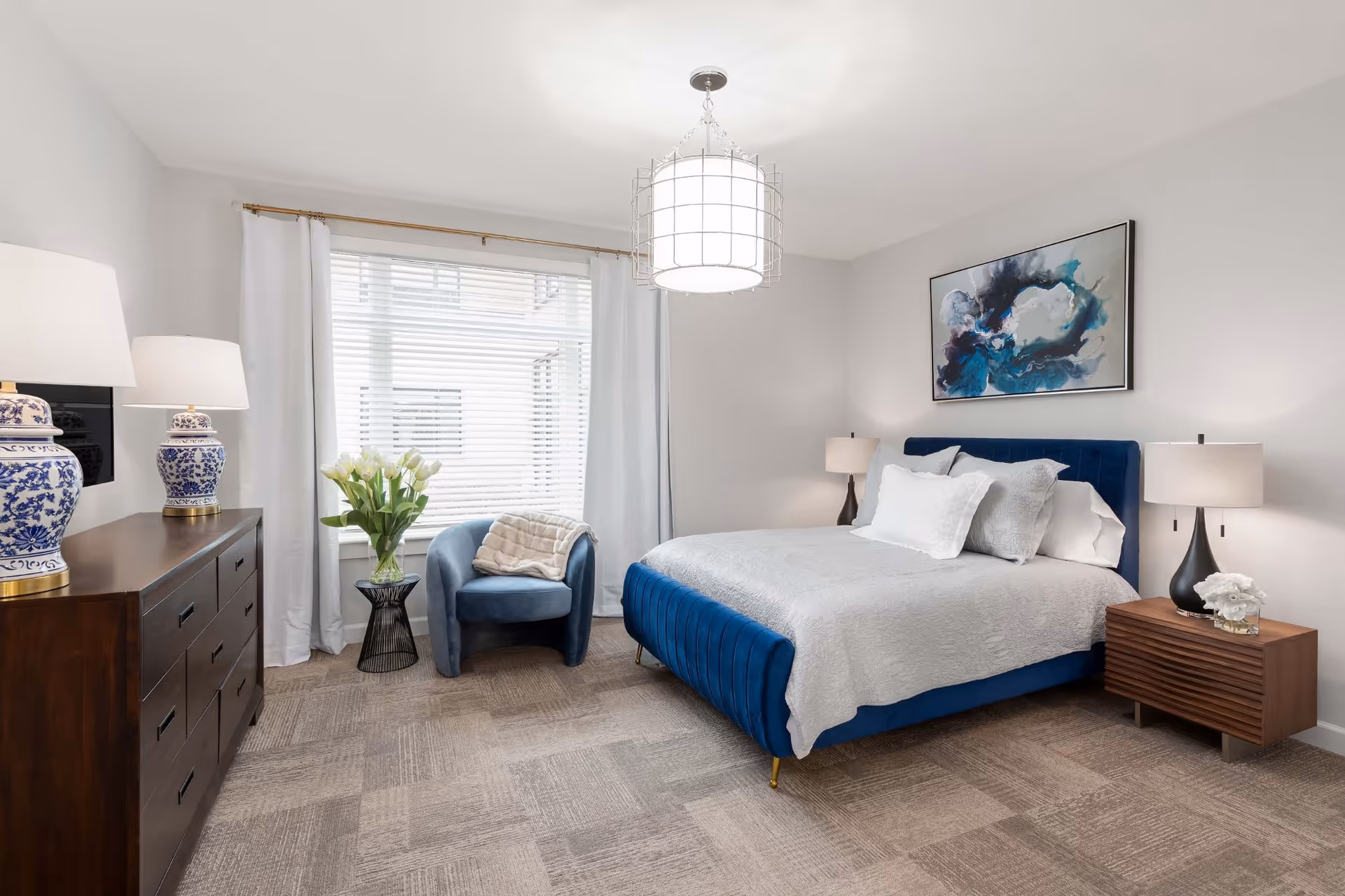 A bright and modern bedroom featuring a blue upholstered bed with white and gray bedding, two bedside tables with lamps, a wooden dresser with two blue and white decorative lamps, a blue armchair with a beige throw blanket, a small black side table with a vase of white tulips, a large window with white blinds and curtains, and an abstract blue and black painting on the wall.