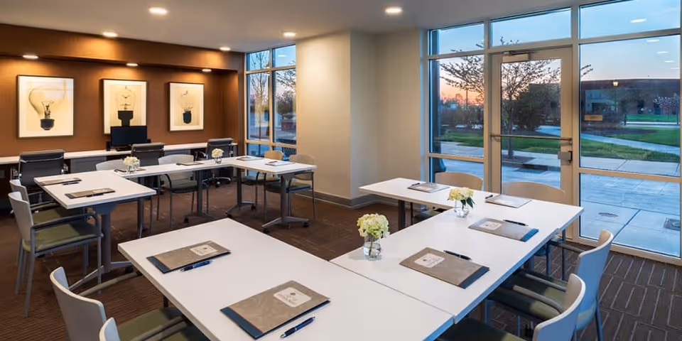 A modern meeting or conference room with white tables arranged in a U-shape, each table set with a folder and pen. The room has large windows letting in natural light and a view of the outdoors. The walls are decorated with framed pictures of light bulbs, and there are several chairs around the tables.