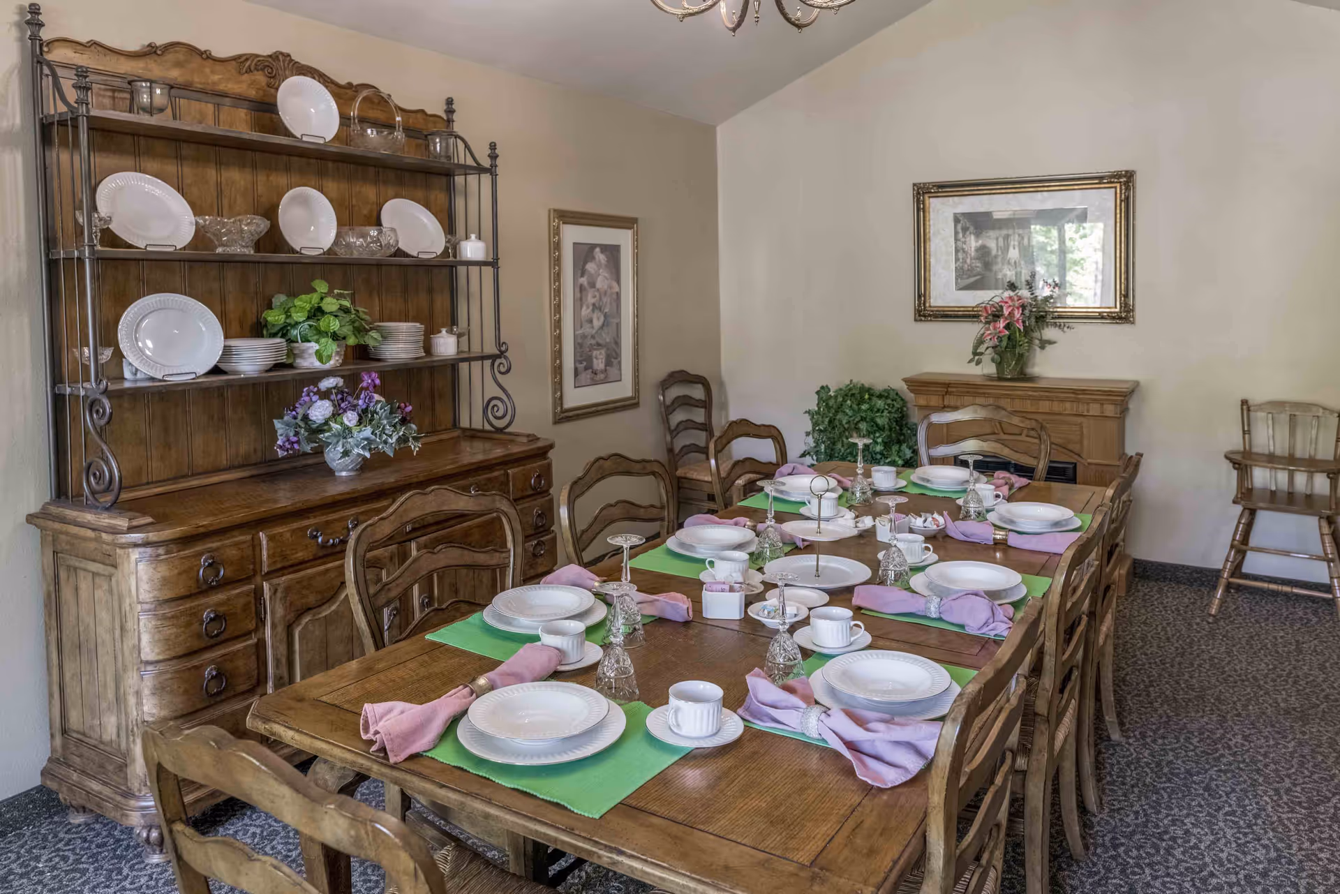 A dining room with a long wooden table set for a meal, wooden chairs, a hutch displaying dishes, and floral decorations.