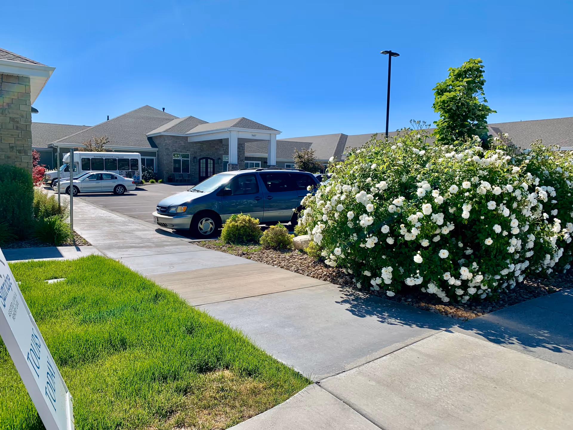 Exterior view of Sunridge Assisted Living and Memory Care facility on a sunny day, showing the building entrance, parked cars, a white flowering bush, and a clear blue sky.