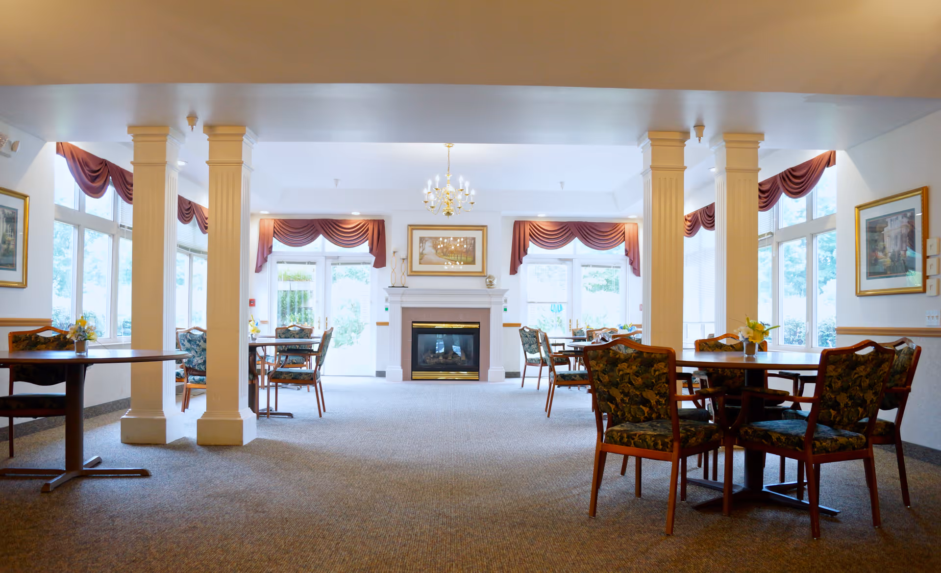 A bright and spacious common area with multiple round tables and chairs arranged for seating. The room features large windows with red valance curtains, white walls, and decorative columns. A fireplace with a framed picture above it is centered on the far wall, and a chandelier hangs from the ceiling.