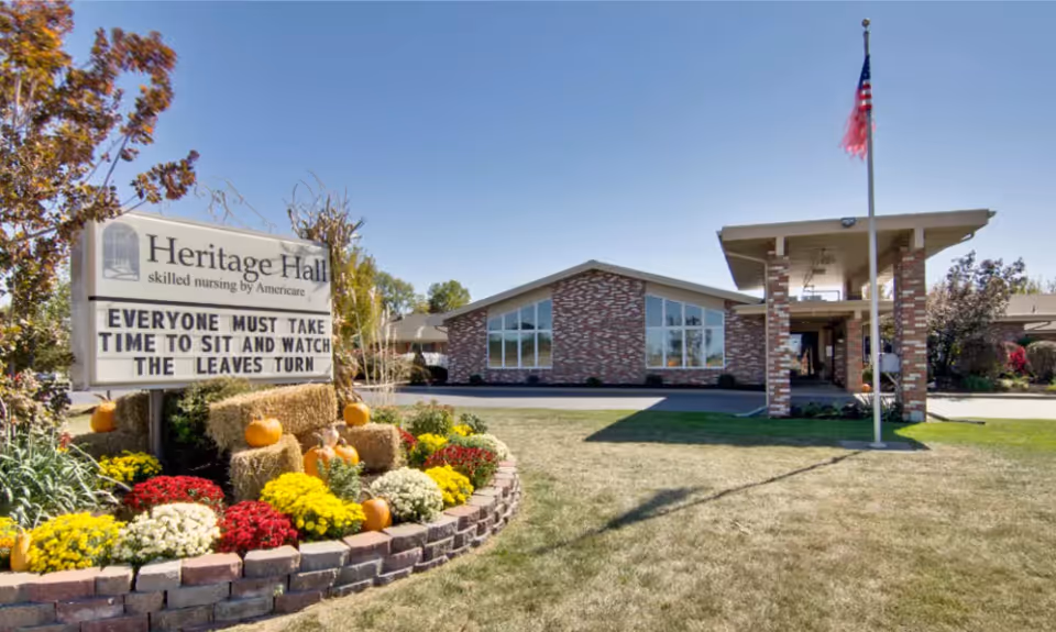 Exterior view of Heritage Hall skilled nursing facility with a brick building, a covered entrance supported by brick pillars, an American flag on a flagpole, and a landscaped area with colorful flowers, pumpkins, and hay bales. A sign in front reads 'EVERYONE MUST TAKE TIME TO SIT AND WATCH THE LEAVES TURN.'