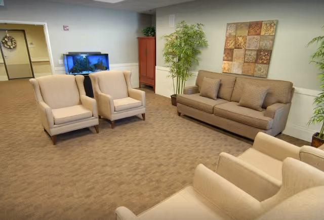 A spacious living room area in an assisted living facility featuring a brown sofa with three cushions, four beige armchairs, a large potted plant, a red cabinet, and a fish tank against the wall. The walls are light-colored with a decorative square wall art piece above the sofa.