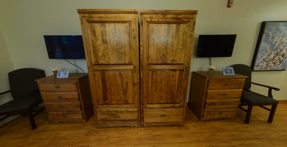 A room with two wooden dressers and a large wooden wardrobe in the center. On each side of the wardrobe, there is a small wooden dresser with three drawers, each topped with a small vase of flowers and a framed photo. Two dark upholstered chairs are placed beside each dresser. Two flat-screen TVs are mounted on the wall behind the dressers. A floral painting is visible on the right side of the image.