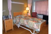 A single bed with a floral patterned bedspread in a room with wooden flooring. Next to the bed is a wooden nightstand with a small stereo system and a lamp. The room has a window with pink curtains and a red armchair beside it.