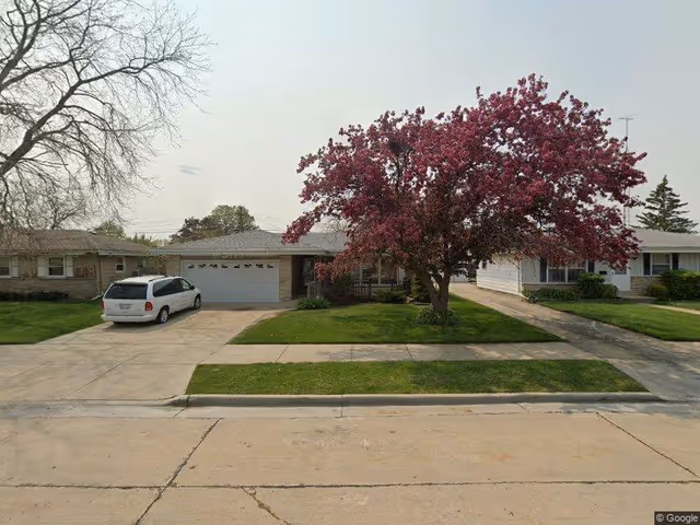 A single-story residential house with a two-car garage and a driveway with a white minivan parked on it. There is a large tree with pink blossoms in the front yard, surrounded by green grass. The house is located in a suburban neighborhood with similar houses on either side.