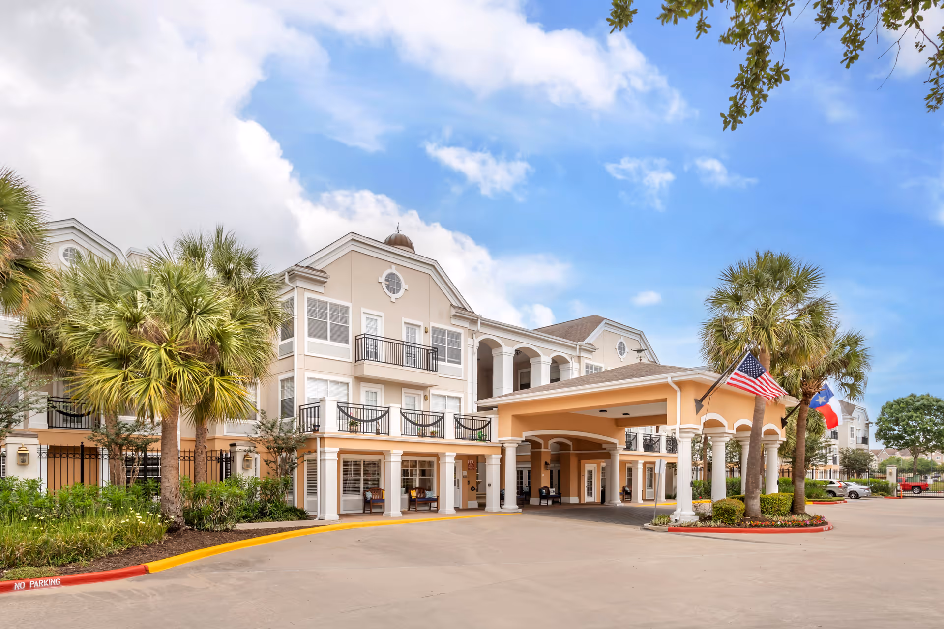 Exterior view of Brookdale Clear Lake senior living facility showing a large beige and white building with multiple windows, balconies, and a covered entrance. There are palm trees and flags, including the American flag and Texas state flag, near the entrance. The sky is partly cloudy with blue patches.