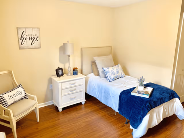 A cozy bedroom with a single bed made with white and blue bedding, a small white nightstand with a lamp, clock, and plant, a beige chair with a 'family' pillow, and a wall decoration that says 'So good to be home'. The room has wooden flooring and light yellow walls.