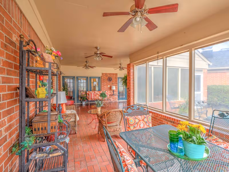 Enclosed sunroom-style patio with brick walls, ceiling fans, wicker and metal dining furniture, colorful cushions, and potted plants.