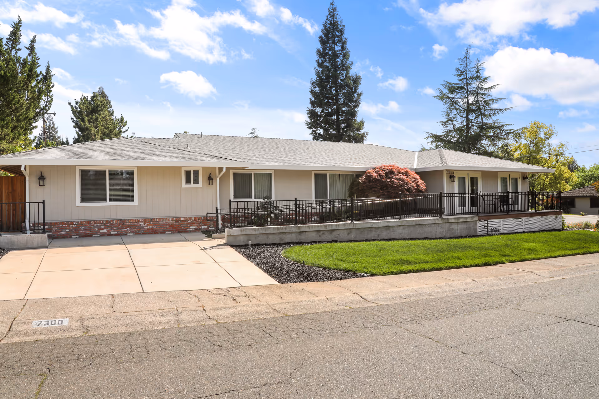Single-story residential building with beige siding and a gray shingled roof, featuring several windows and a ramp with black railings leading to the entrance. The front yard has a well-maintained green lawn and a small tree with red leaves. The sky is partly cloudy with blue patches.