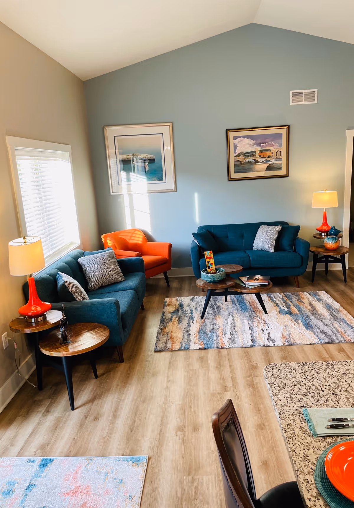 A cozy living room with two teal sofas, an orange armchair, two wooden side tables with orange lamps, a wooden coffee table with books and a decorative bowl, and two framed pictures on a light blue wall. The room has light wood flooring and two colorful area rugs.