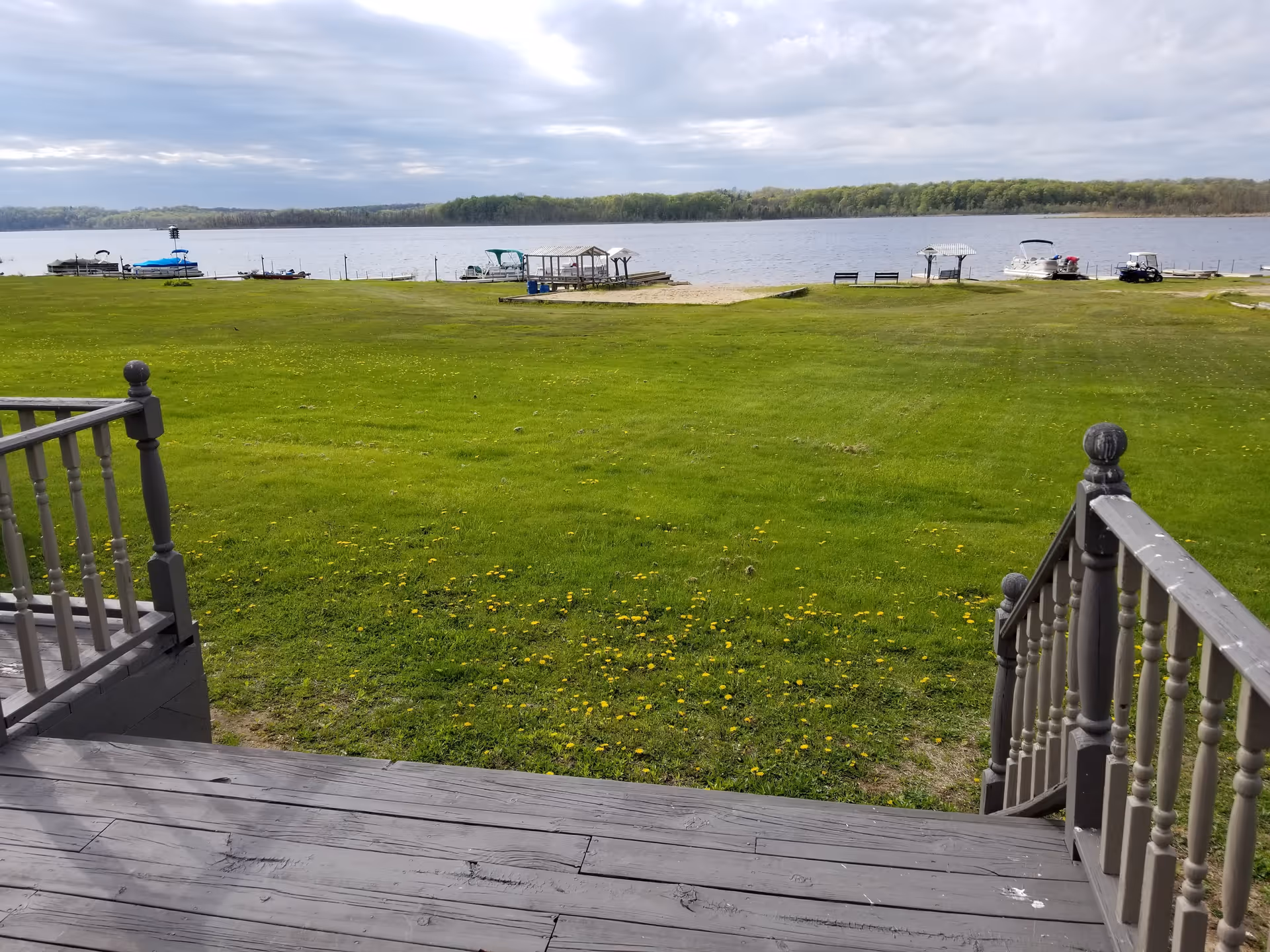 View from wooden steps leading down to a large green lawn with scattered yellow flowers, overlooking a lake with several boats docked along the shore and a tree-lined horizon under a cloudy sky.