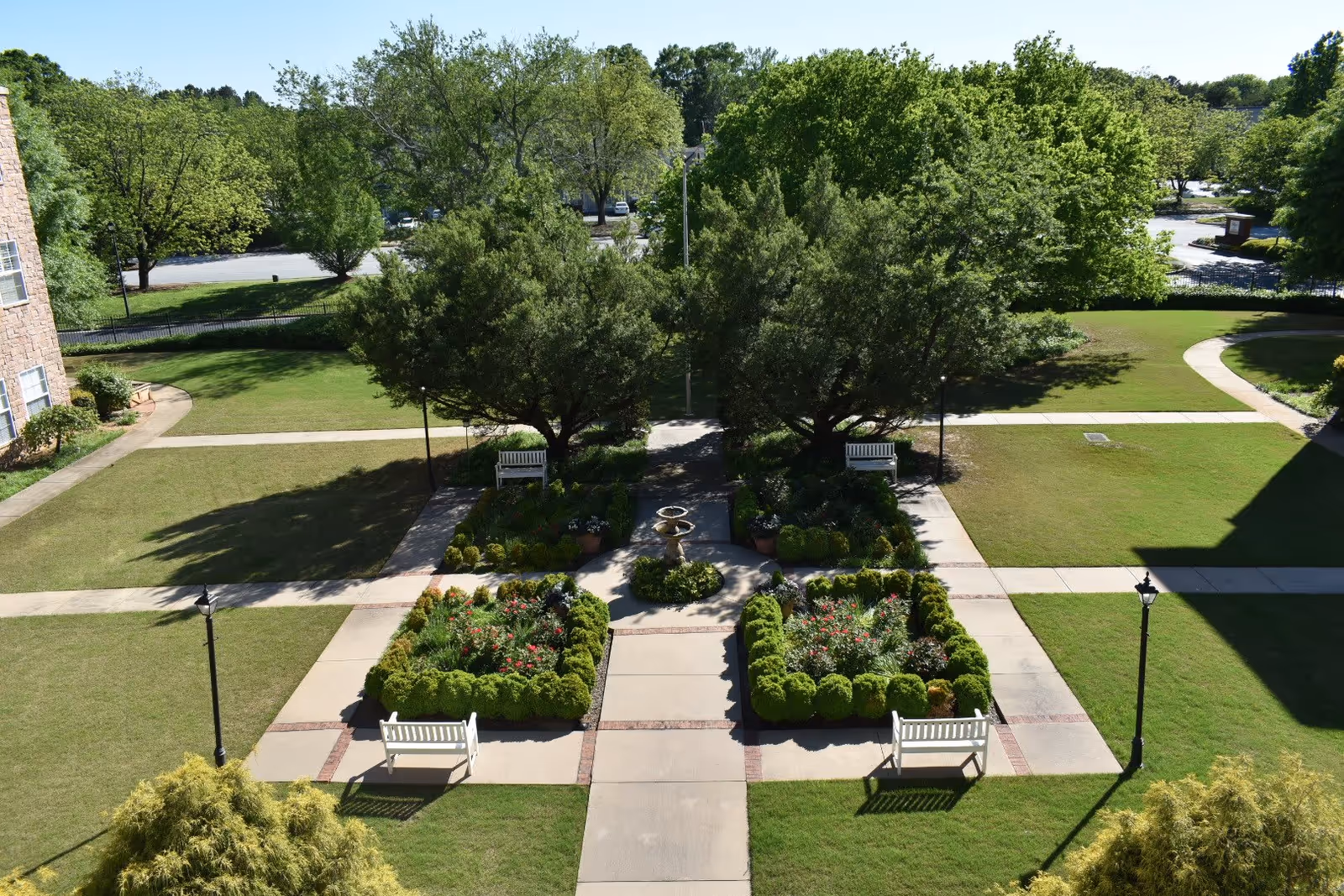 A well-maintained outdoor garden area with neatly trimmed bushes, flower beds, and four white benches arranged around a central paved walkway. Two large trees provide shade near the center, and there are lamp posts along the paths. The garden is surrounded by green lawns and trees in the background under a clear sky.