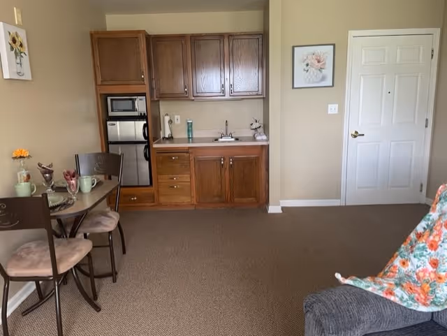 A small kitchenette area with wooden cabinets, a microwave, and a mini refrigerator. To the left, there is a small dining table with two chairs, set with cups and a small flower vase. On the right side, part of a gray upholstered chair with a floral blanket is visible. The walls are beige with a framed floral picture and a white door.