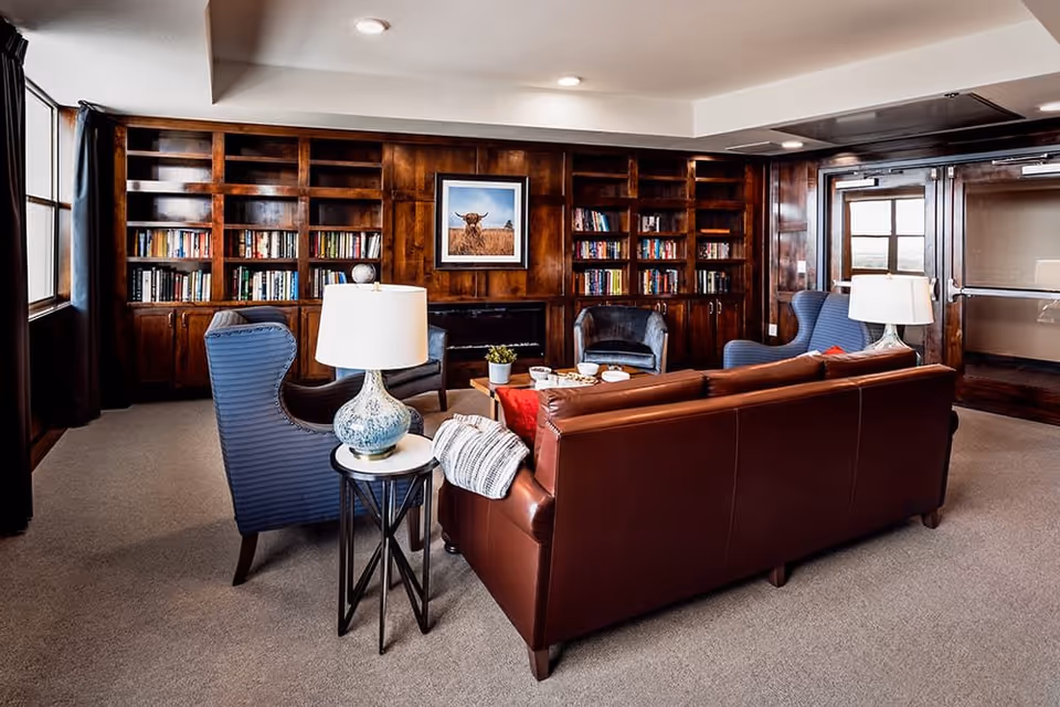 A cozy living room with a brown leather sofa, two blue armchairs, a small round side table with a decorative lamp, and built-in wooden bookshelves filled with books. A framed picture of a cow hangs above a fireplace in the background. The room has carpeted flooring and large windows with dark curtains.