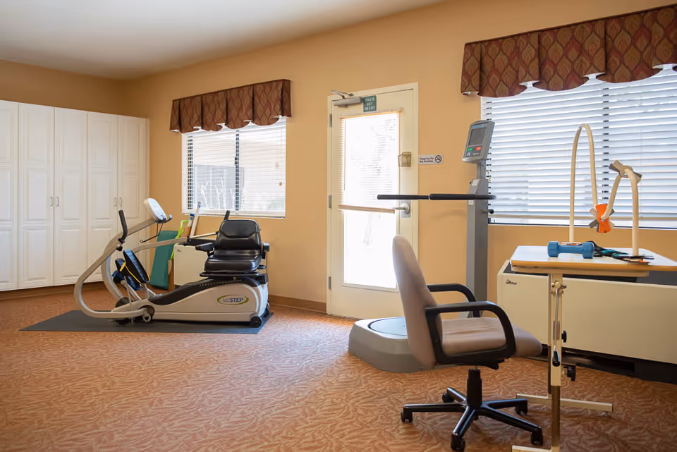 A well-lit exercise room with a recumbent exercise bike, a vibration plate machine, a chair, and a table with exercise equipment. The room has beige walls, carpeted floor, two windows with blinds and valances, and a door with a sign that reads 'This is not an exit.'