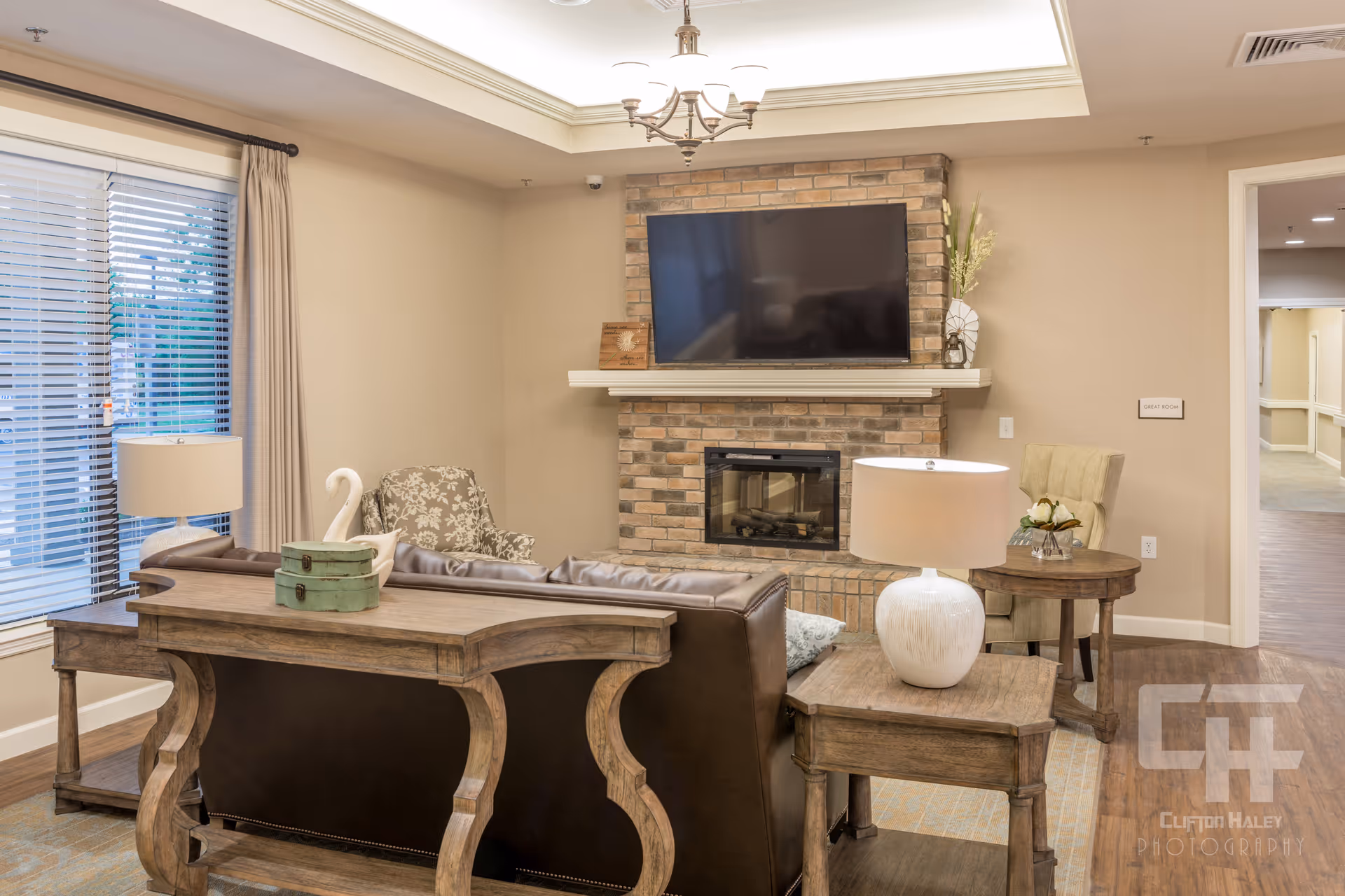 A cozy living room area in a senior living facility featuring a brown leather sofa, wooden side tables with lamps, a patterned armchair, a brick fireplace with a mounted flat-screen TV above it, and a window with blinds and curtains. The room has beige walls, wooden flooring, and a ceiling light fixture.
