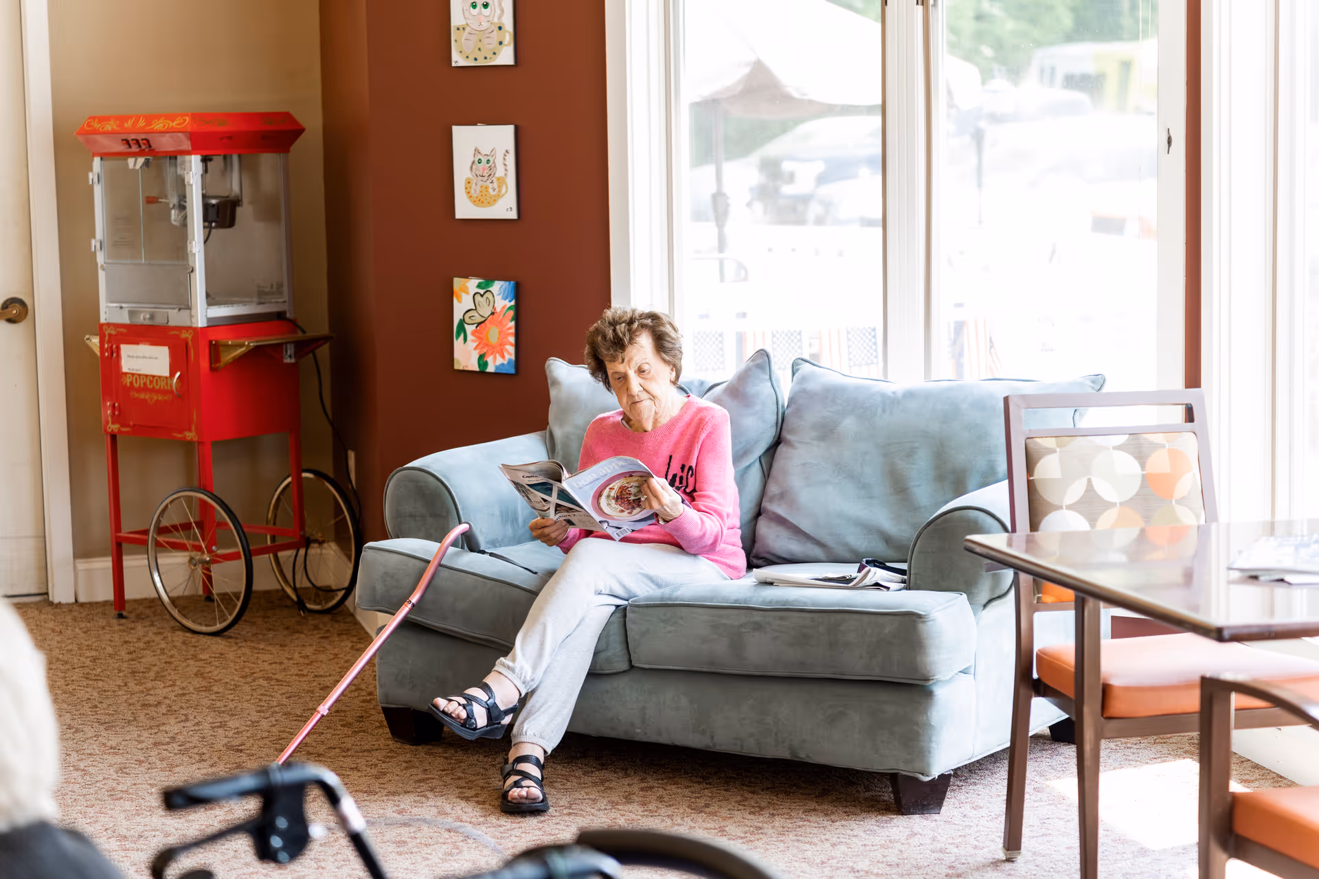 An elderly woman sits on a light blue couch reading a magazine in a bright common room with a popcorn machine and a table nearby.
