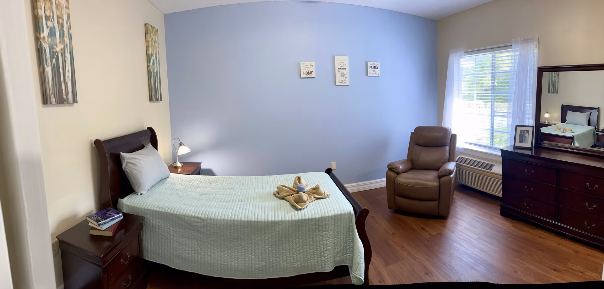 A neatly arranged bedroom with a single bed covered in a light green quilt, a pillow, and a towel folded into a decorative shape on top. There is a wooden nightstand with books and a lamp beside the bed. On the opposite side, there is a comfortable brown recliner chair near a window with white blinds and sheer curtains. A wooden dresser with a large mirror and a framed photo is also visible. The walls are painted light blue and beige, decorated with three small framed quotes and two paintings of trees. The floor is wooden.