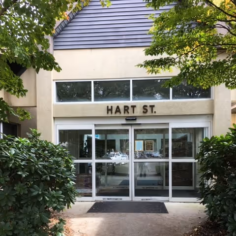 Exterior entrance of a building with glass sliding doors under a sign reading 'HART ST.' framed by shrubs and trees.