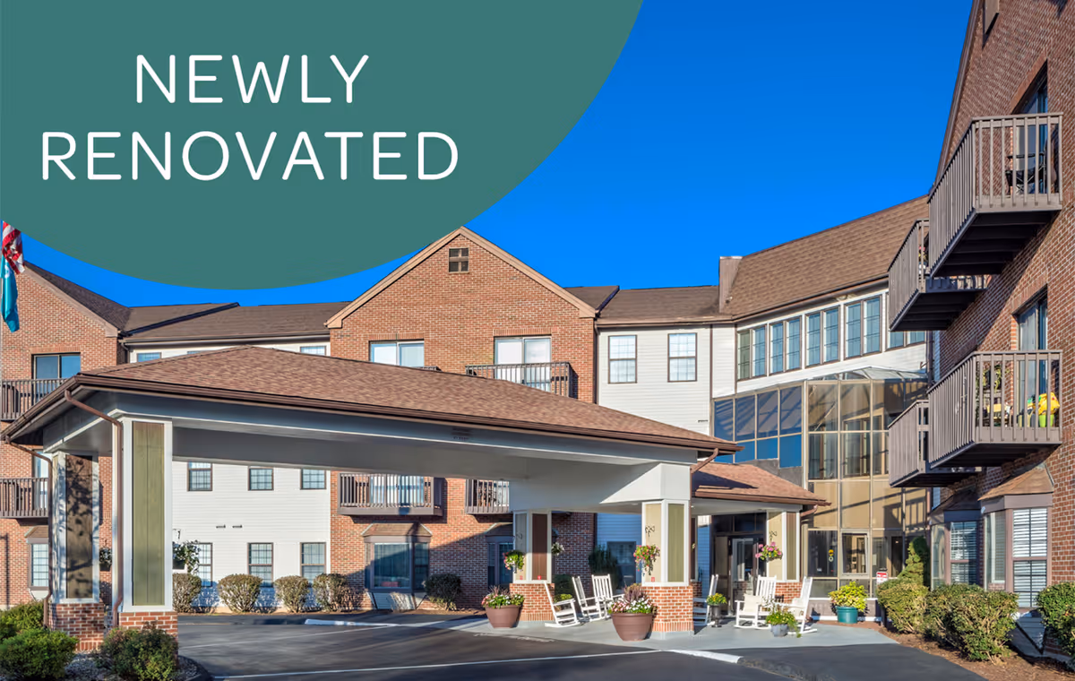 Exterior view of a senior living facility with a covered entrance, brick and siding walls, balconies, and several potted plants and rocking chairs near the entrance under a clear blue sky. Text overlay in the top left corner reads 'NEWLY RENOVATED'.