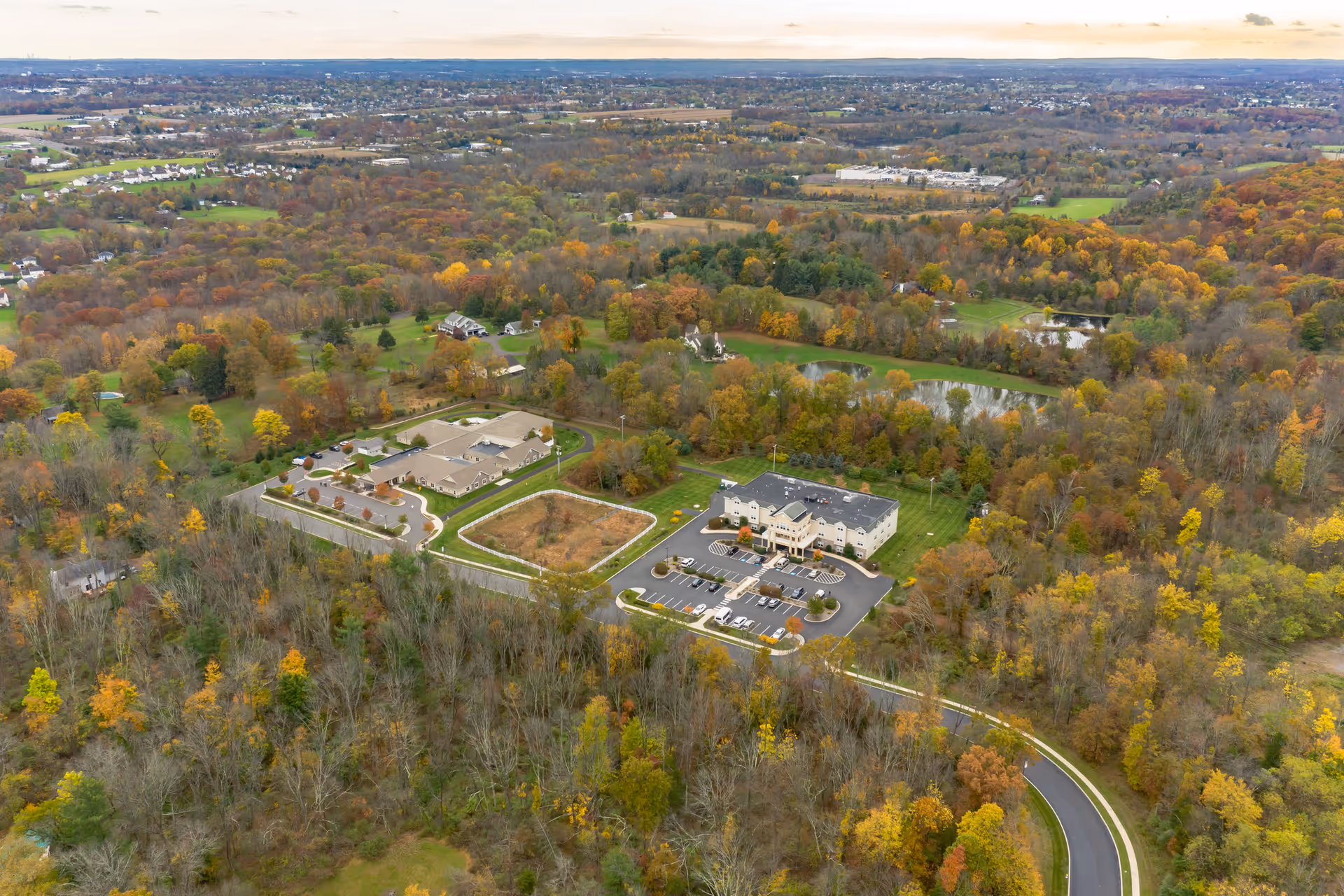 Aerial view of Hidden Meadows On the Ridge facility surrounded by dense trees with autumn foliage. The image shows two main buildings with parking lots, a curved road leading to the facility, and a large area of forest and open land extending into the distance under a cloudy sky.