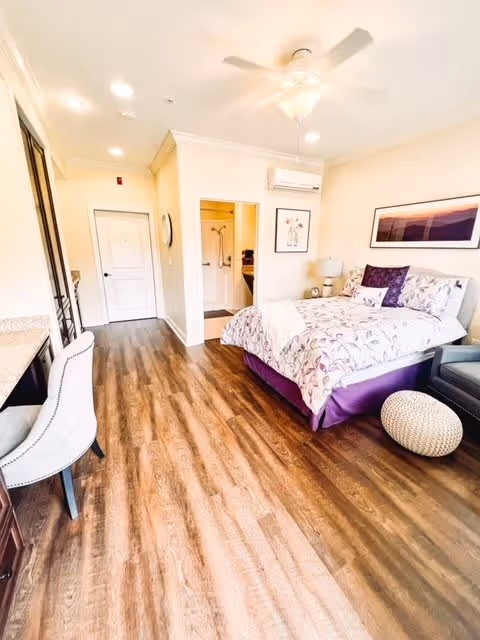 A bright bedroom with wood flooring featuring a bed with floral bedding and purple accents, a gray armchair, a round woven ottoman, a desk with a white chair, a ceiling fan, and an open doorway leading to a bathroom.