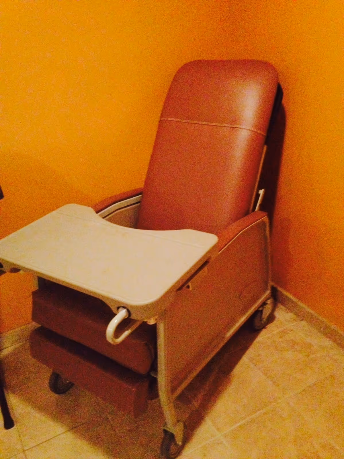 A brown reclining medical chair with a white attached tray table, positioned in a corner with orange walls and tiled floor.