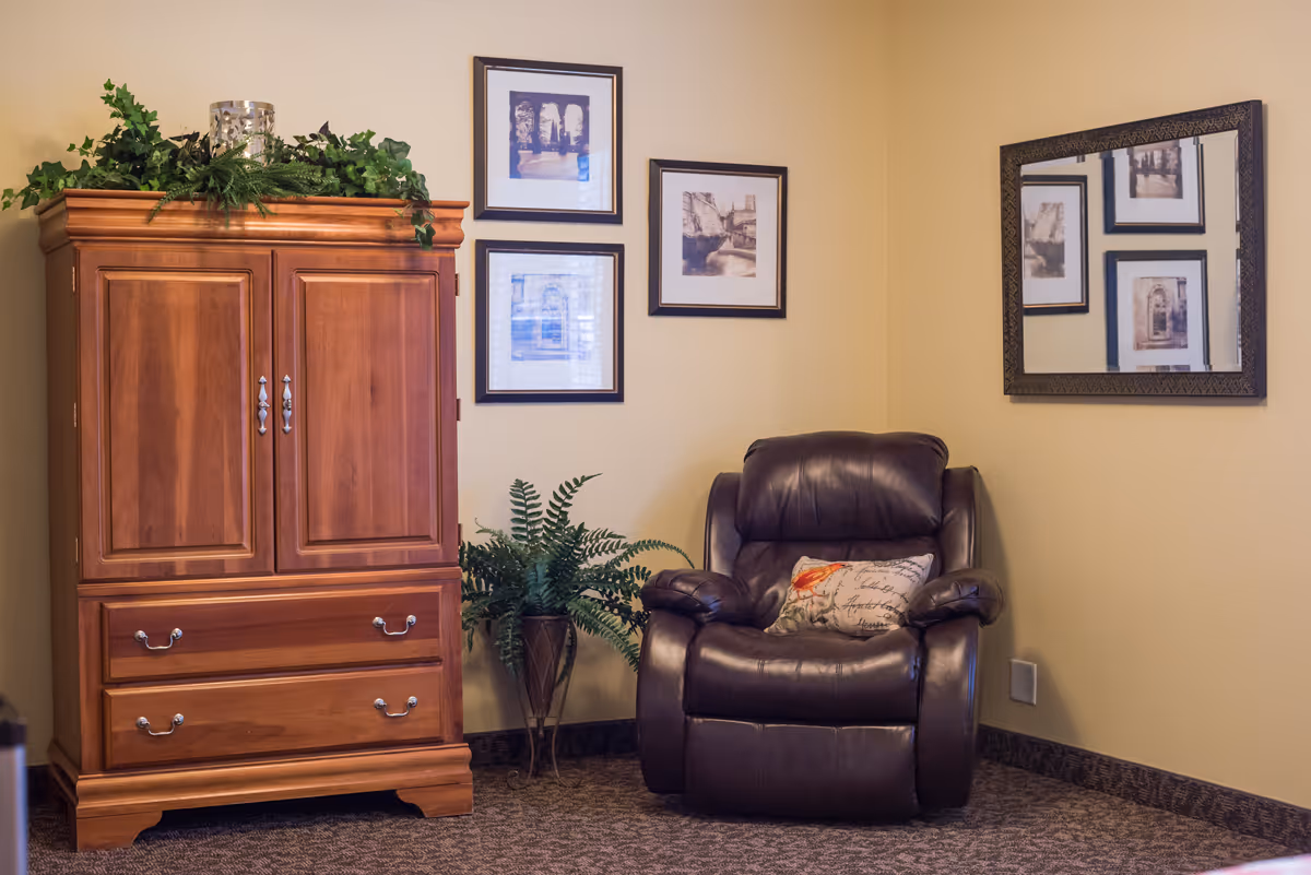 Cozy sitting area with a wooden armoire, potted plant, a brown leather recliner, and framed artwork on beige walls.
