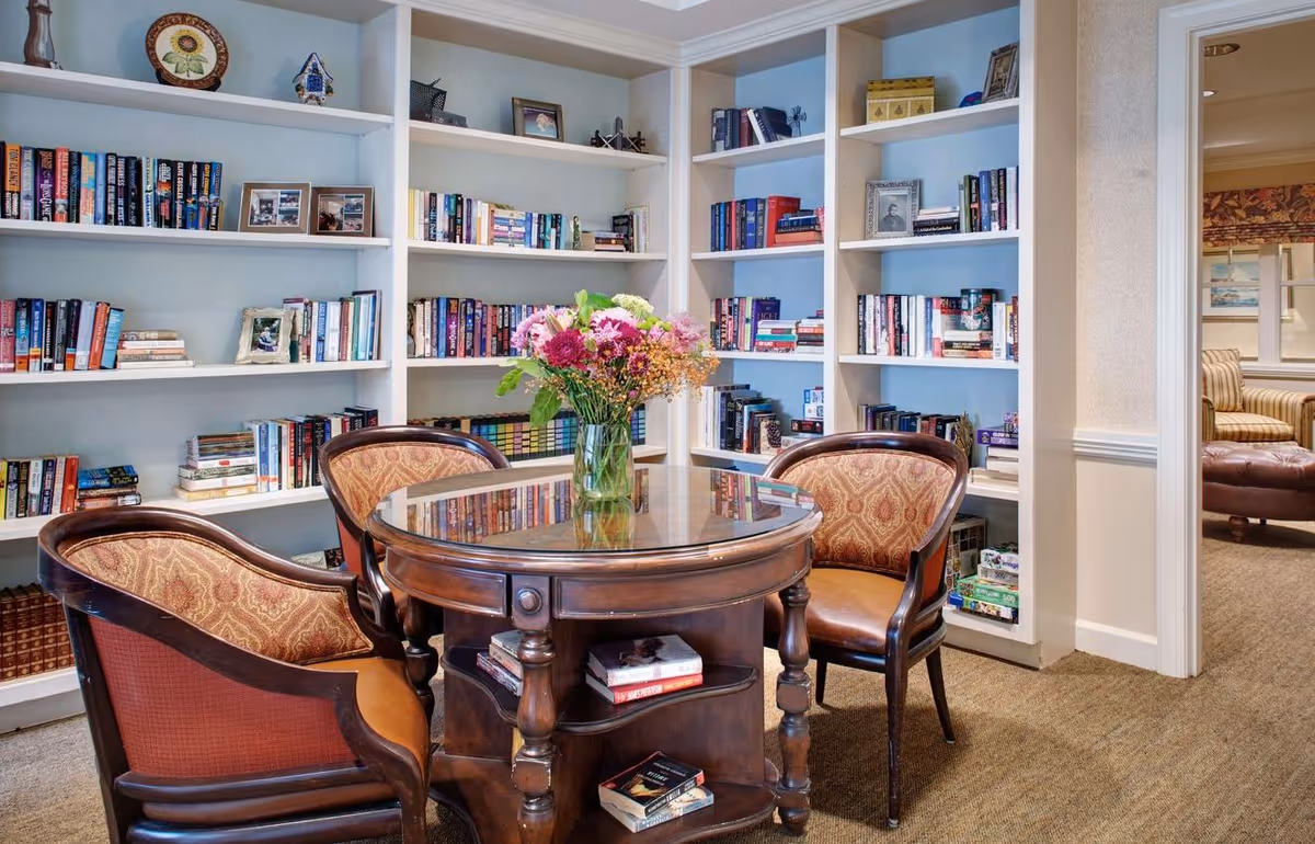 A cozy reading nook with a round wooden table and four upholstered chairs. The table has a glass top and a vase with a bouquet of colorful flowers. Behind the table are built-in white bookshelves filled with books, framed photos, and decorative items. To the right, there is a doorway leading to another room with additional seating.