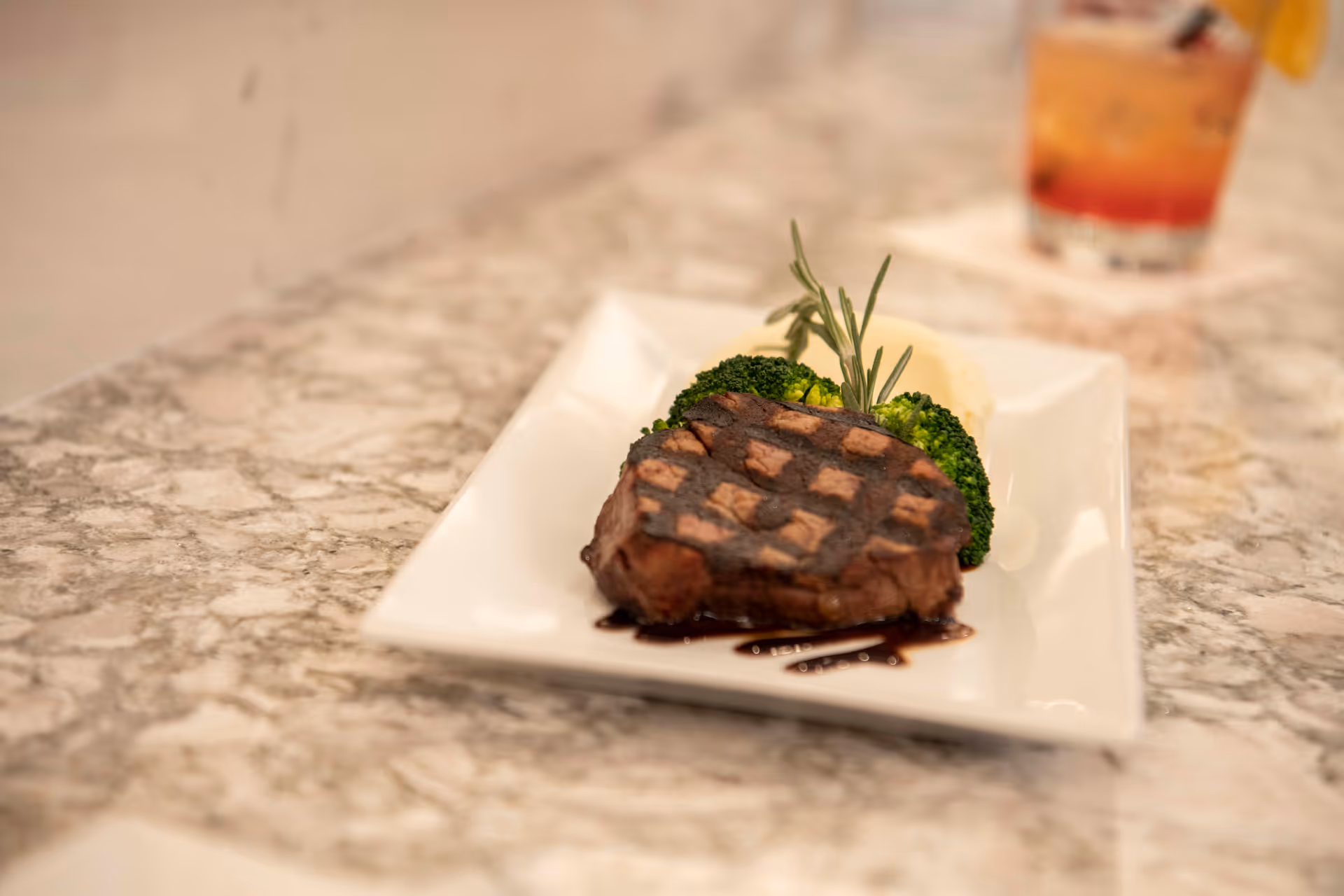 A plated meal featuring a grilled steak with crosshatch grill marks, served with steamed broccoli and mashed potatoes, garnished with a sprig of rosemary on a white rectangular plate on a marble countertop. A blurred drink with a lemon garnish is visible in the background.