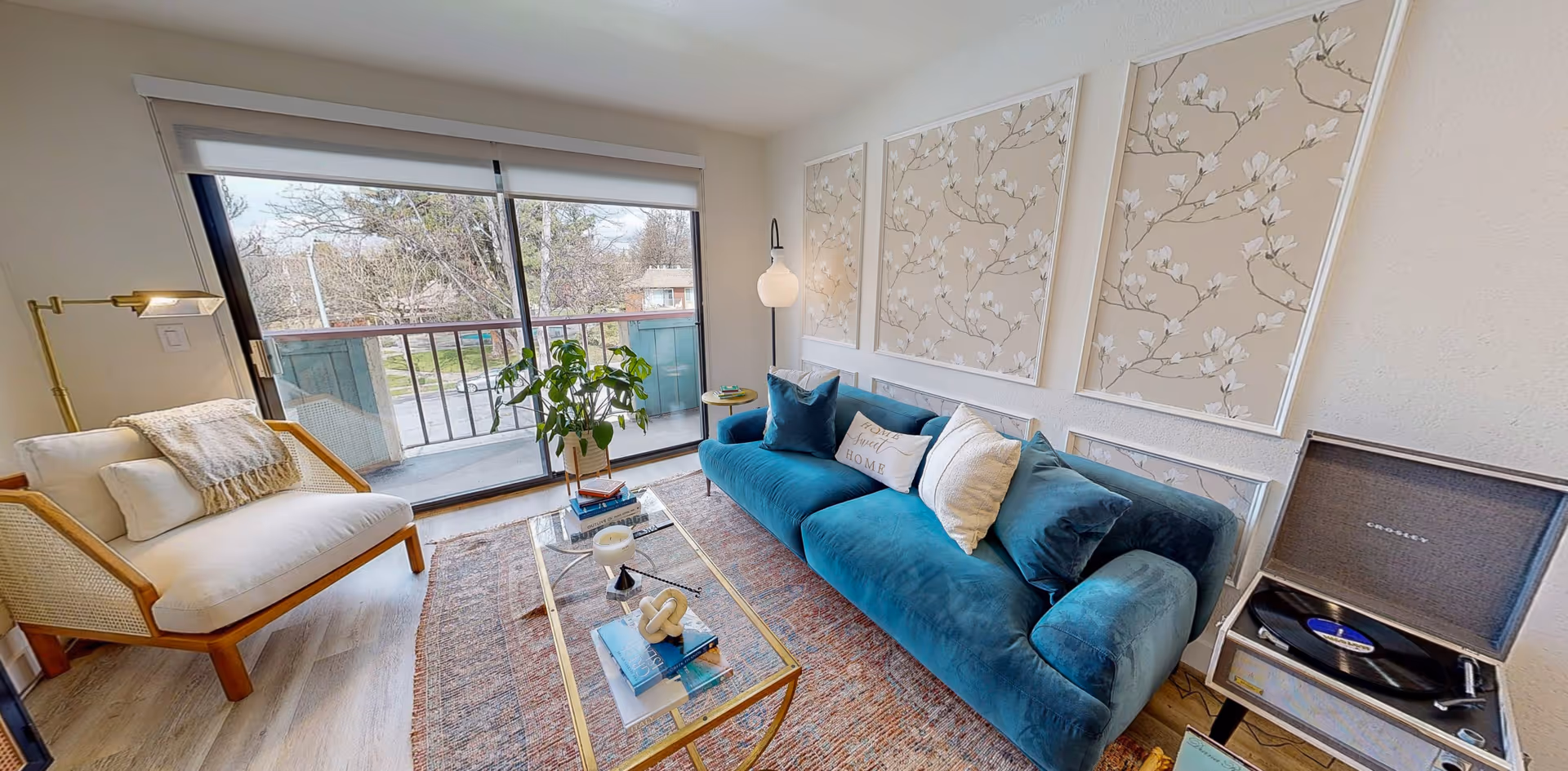 Bright living room with a teal velvet sofa, armchair, glass coffee table, wall art, and a sliding glass door to a balcony.