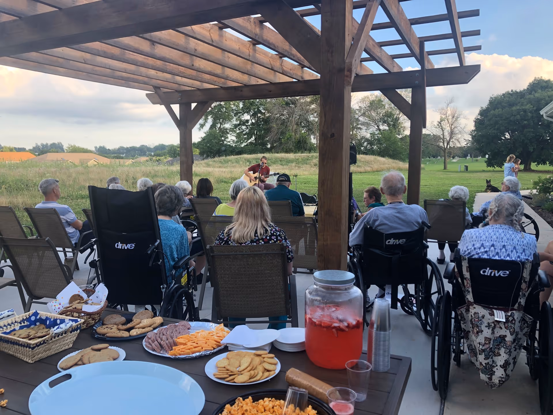 Residents seated under a wooden pergola, many in wheelchairs, watching a musician perform while snacks and a drink dispenser sit on a table in the foreground.