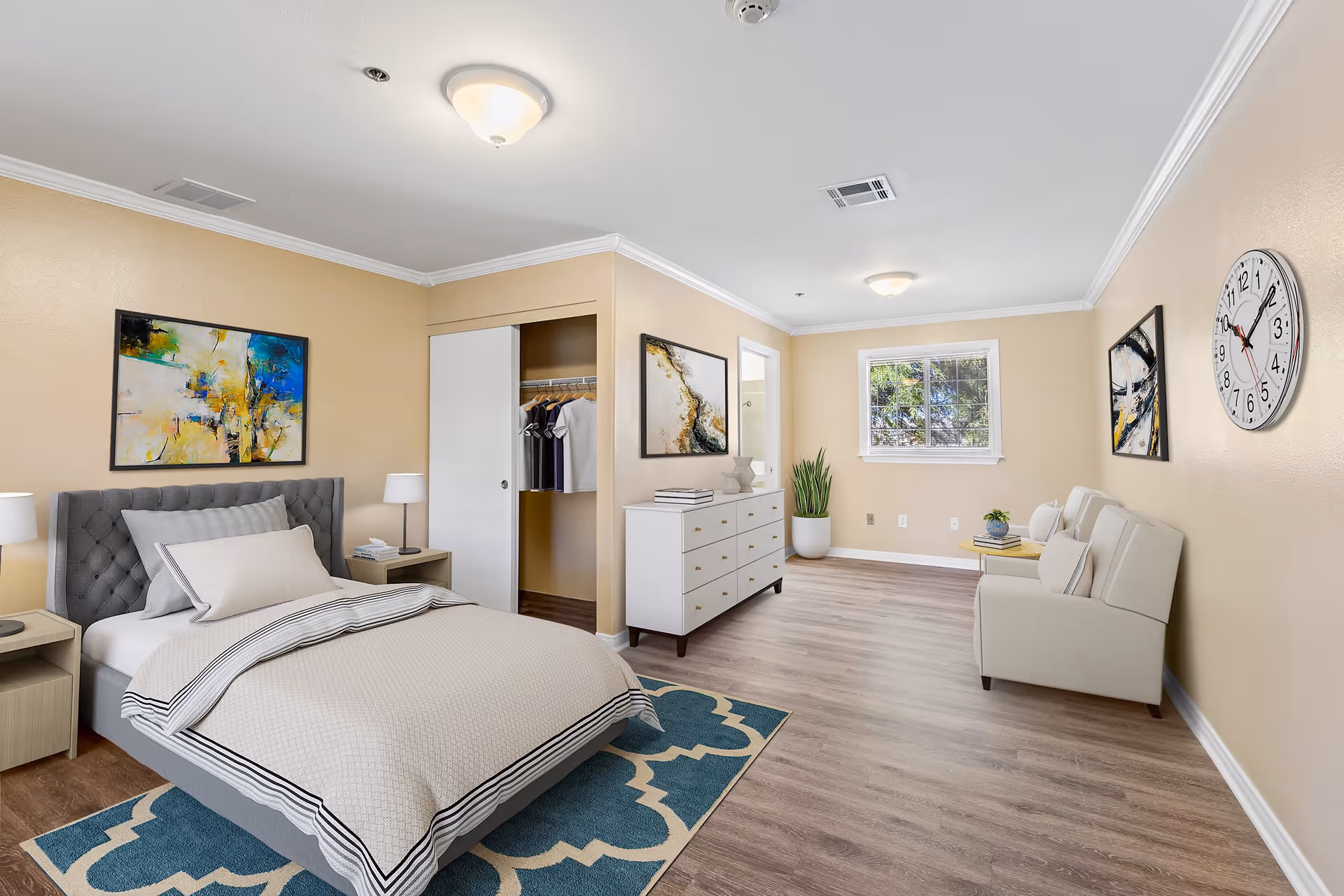 A spacious bedroom with beige walls and wood flooring. The room features a gray upholstered bed with beige bedding, two bedside tables with lamps, an open closet with hanging clothes, a white dresser with decorative items, a large window letting in natural light, a beige loveseat with pillows, a round side table with a plant, a large wall clock, and abstract paintings on the walls.